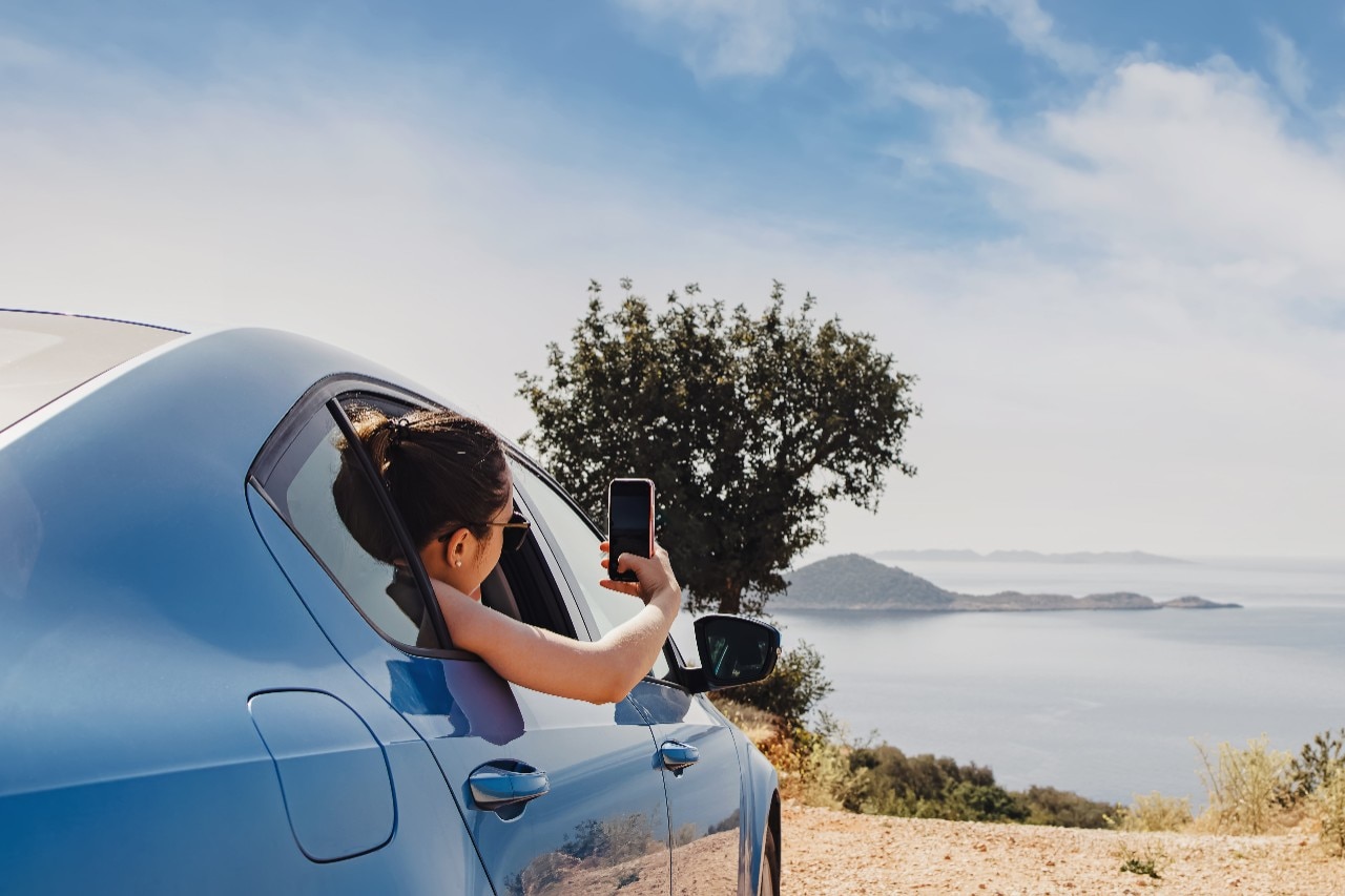 Young woman taking picture with smartphone from open window of a car stopped roadside with a beautiful view to the coast